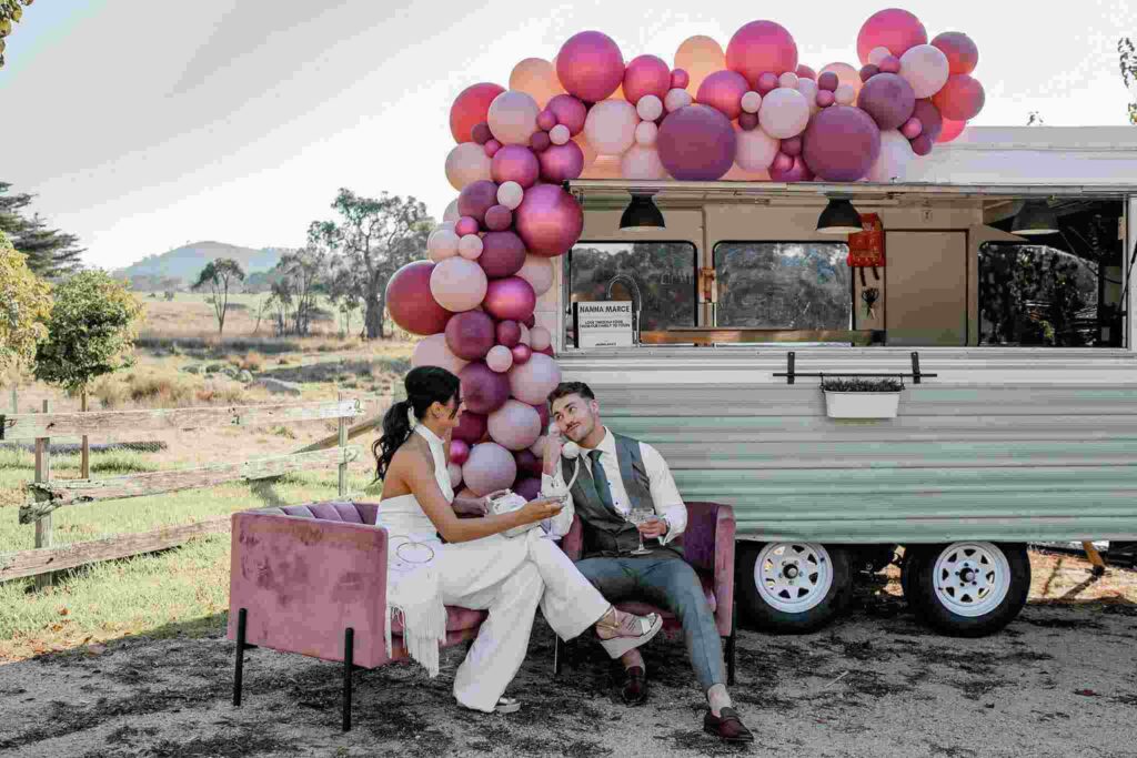 A couple in front of a van, sitting under a balloon arch and talking on a phone