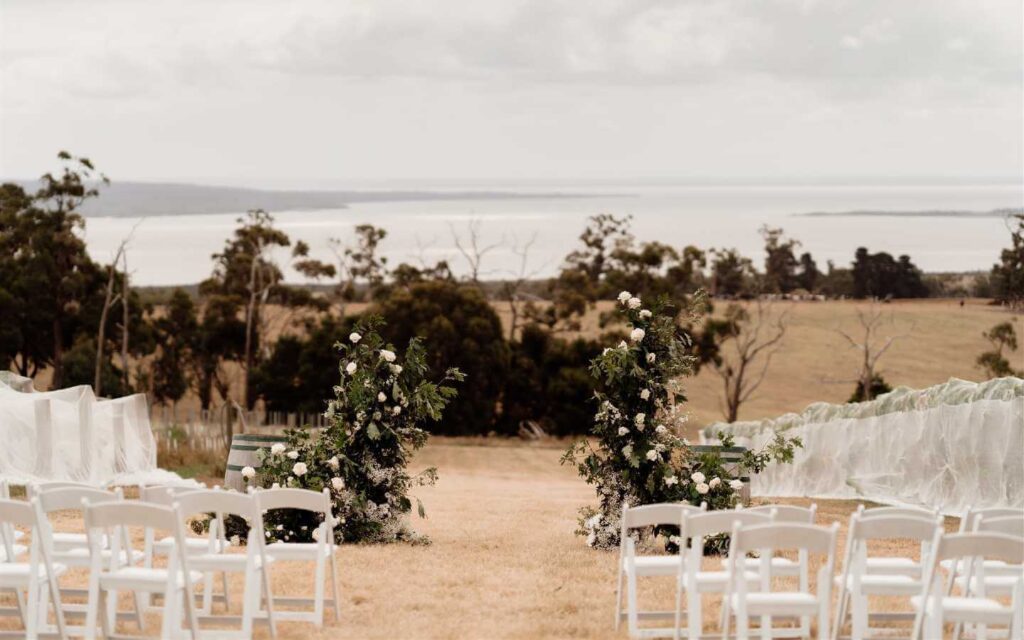 Flowers form an arbour at the end of an aisle using white flowers with a rural paddock as the background
