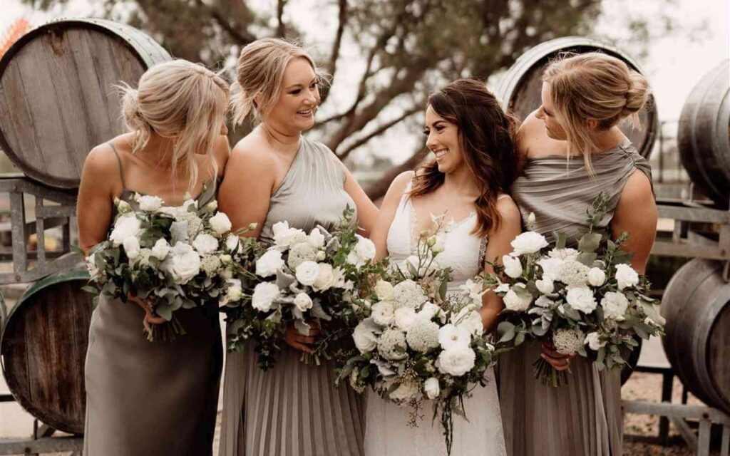 Bride stands with bridal party in champagne colour dresses holding white bouquets in front of wine barrels