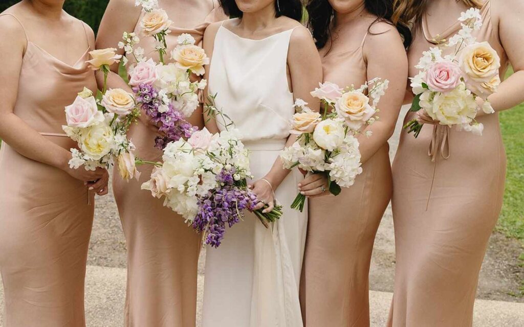 Bride holds wedding bouquet with purple and cream flowers surrounded by bridesmaids wearing champagne colour and holding bouquets of pink roses and white flowers