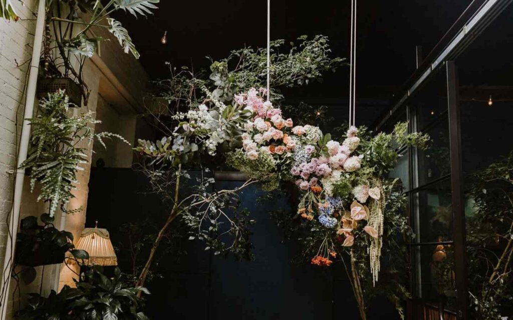 Grand floral feature piece suspended overhead in dimly lit room with greenery surrounding. The installation has foliage and a varied collection of orange, pink and cream florals