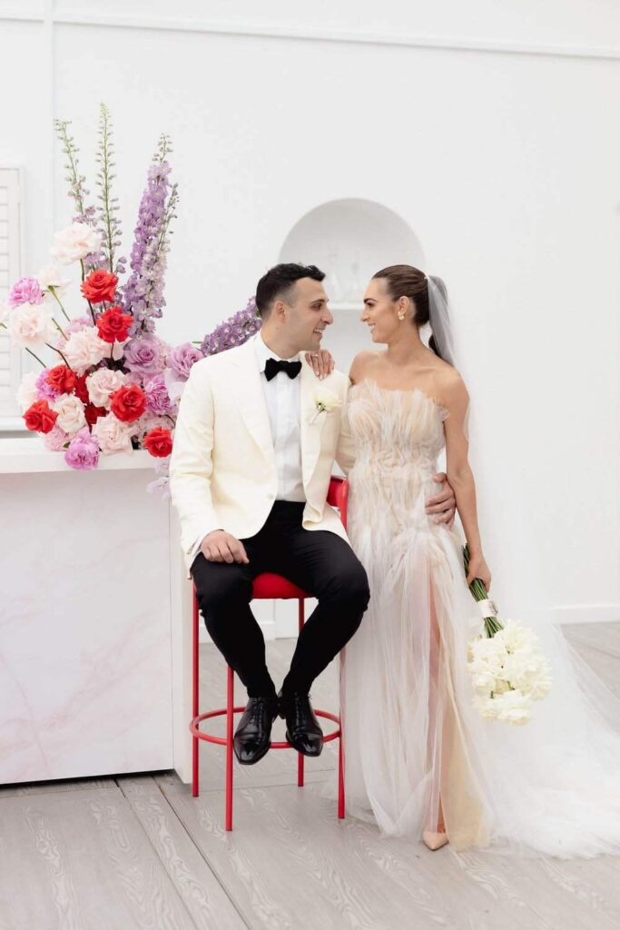Bride and groom stand in white bright room with white bouquet and red purple and baby pink floral arrangement sitting on bar behind them