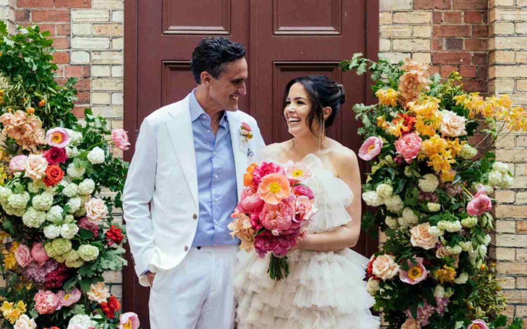 Bride and groom stand in front of a brown door and two pillars of colourful flowers