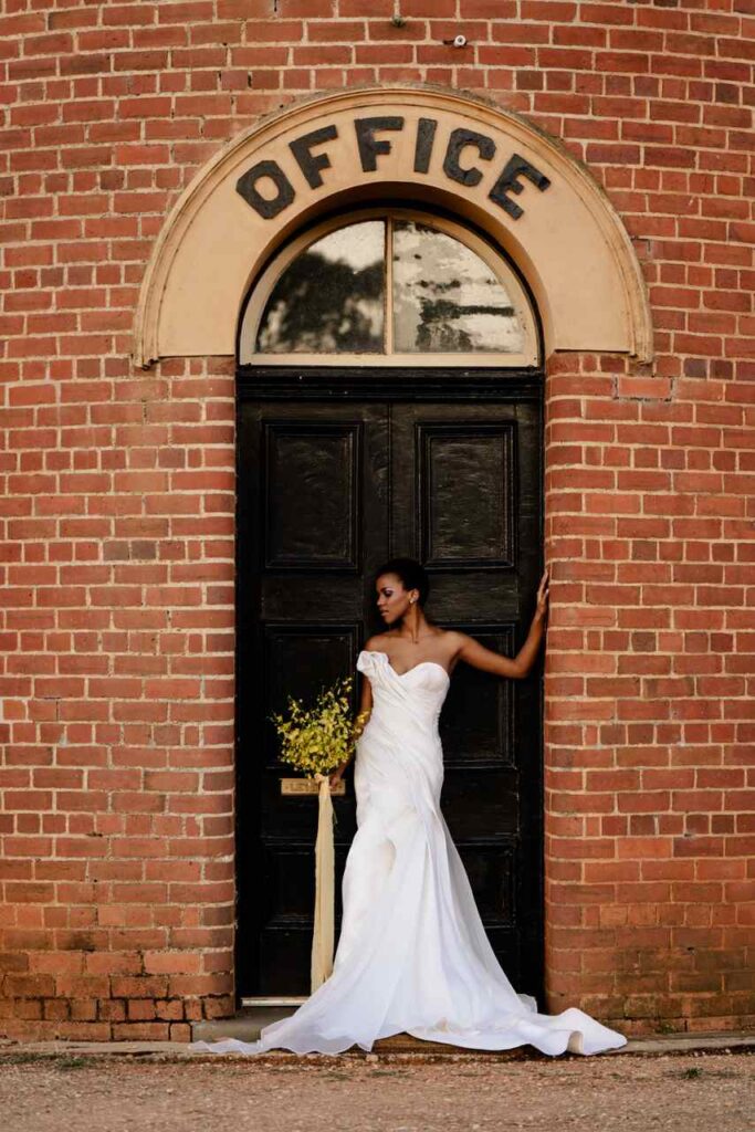 Bride holds wedding bouquet with yellows in front of brick wall that says office above a black door