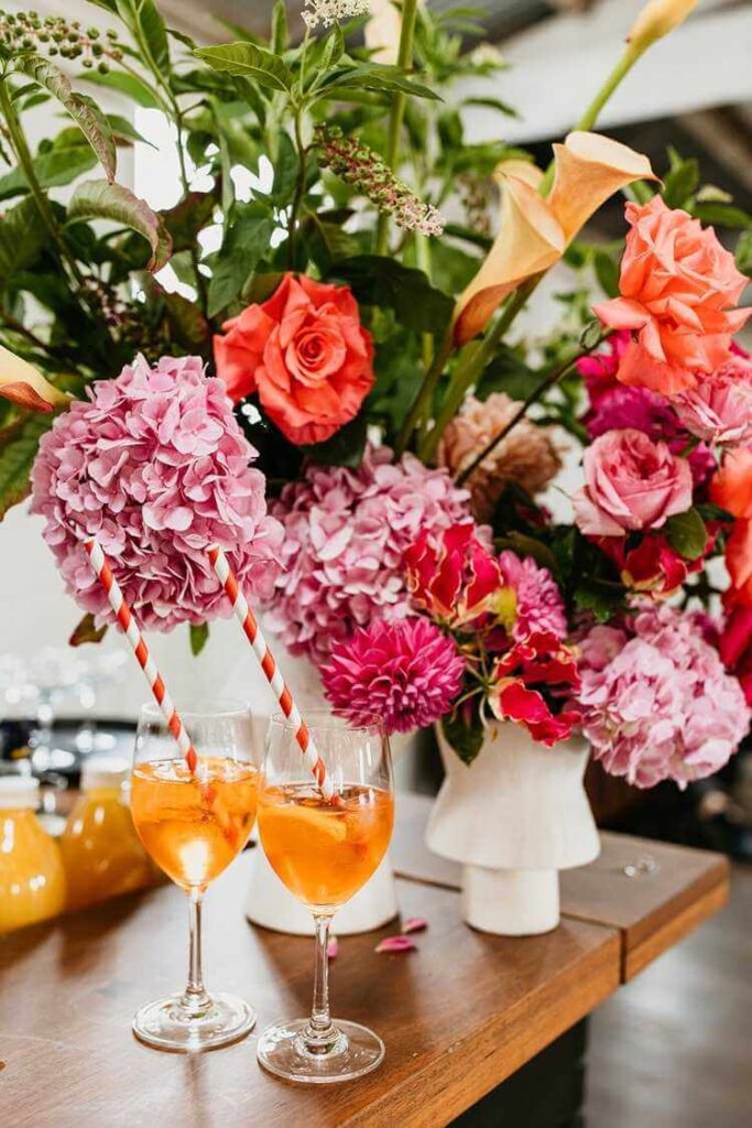 Two aperol spritzes on a table beside a vase of pink and purple flowers