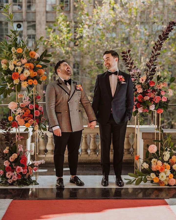 Grooms laughing holding hands surrounded by orange and pink florals