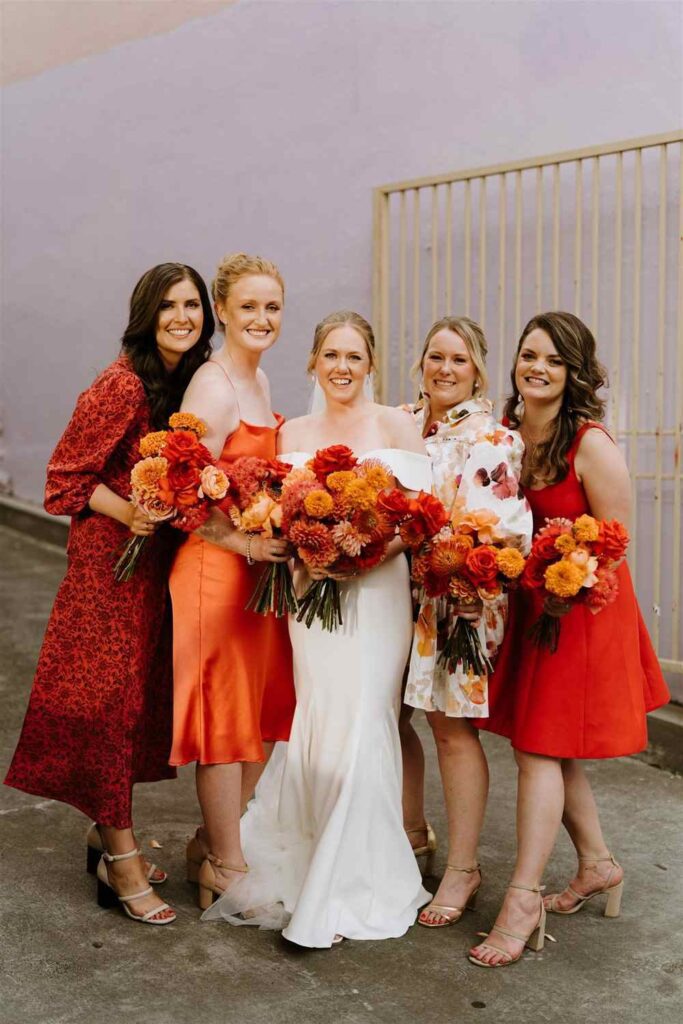 Bride wearing white floor length dress stands with bridesmaids in burnt orange with orange and red bouquets