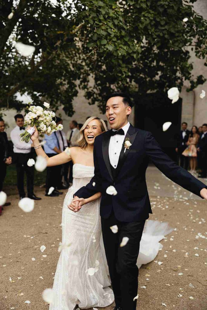 couple leaving ceremony with white rose petals floating in air while smiling at guests