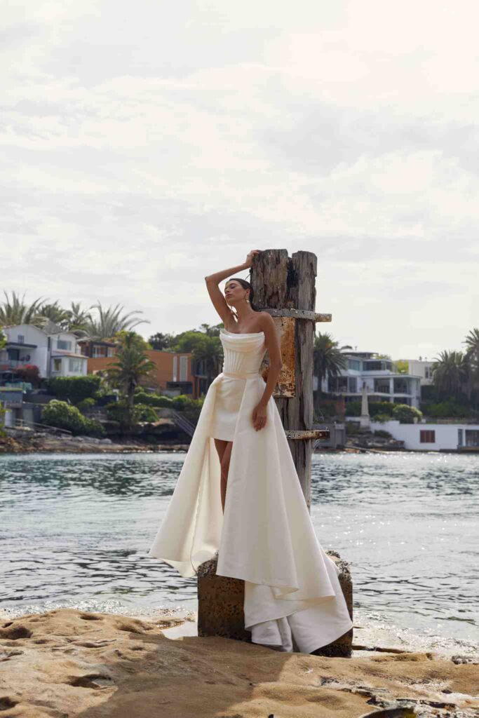 Model wears white silk dress with short 
skirt and long caping sides while standing by water’s edge with houses in the background