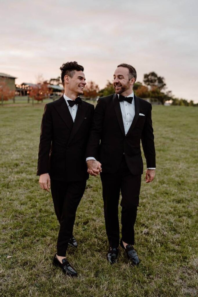 Two grooms walk together holding hands in a field on their wedding day