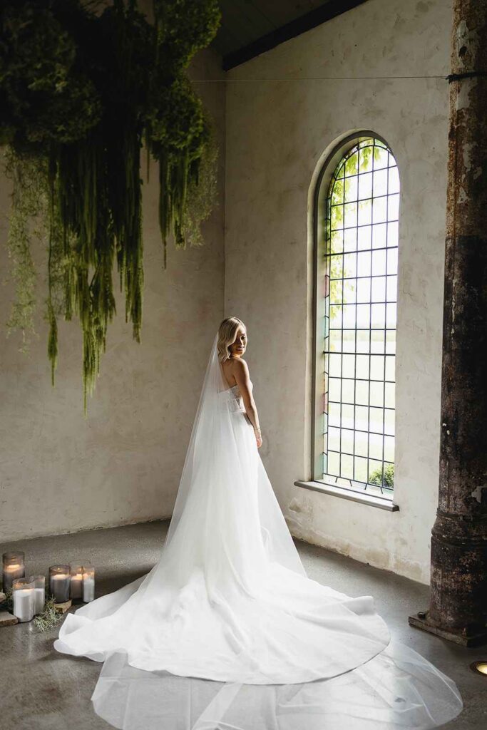 A blushing bride bathes in the sunlight peeking through a window at her wedding ceremony venue.