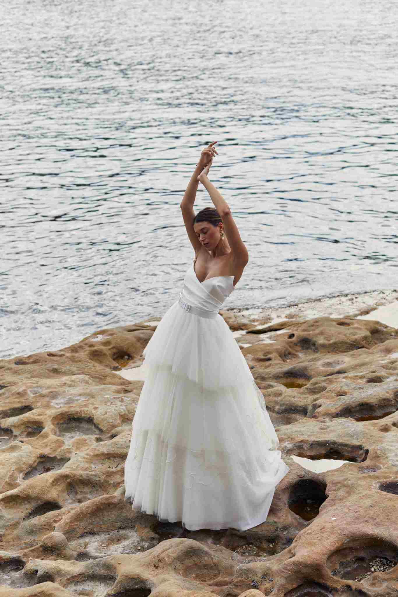 Bride with arms above head standing on top of stone beside the ocean water.