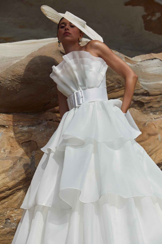 Bride in white hat poses against rocks in wedding dress.