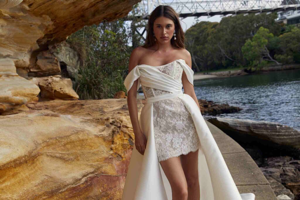 Bride poses in lace and silk wedding gown between stone wall and ocean water.