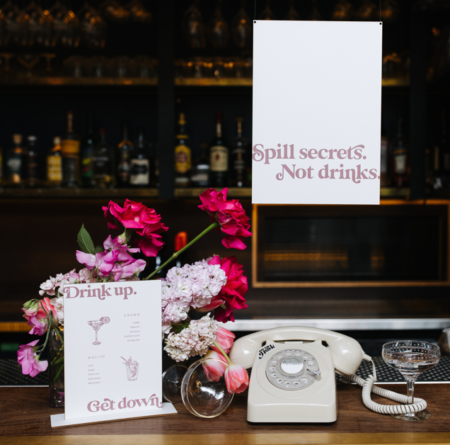 A couple of wedding bar menus on top of a bar surrounded by flowers and a phone.